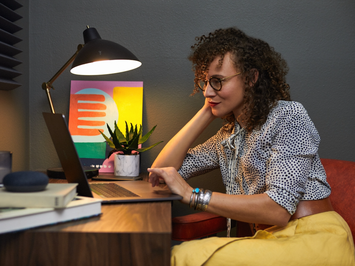 An adult working at a sunlit home desk with a laptop, books, and a plant nearby