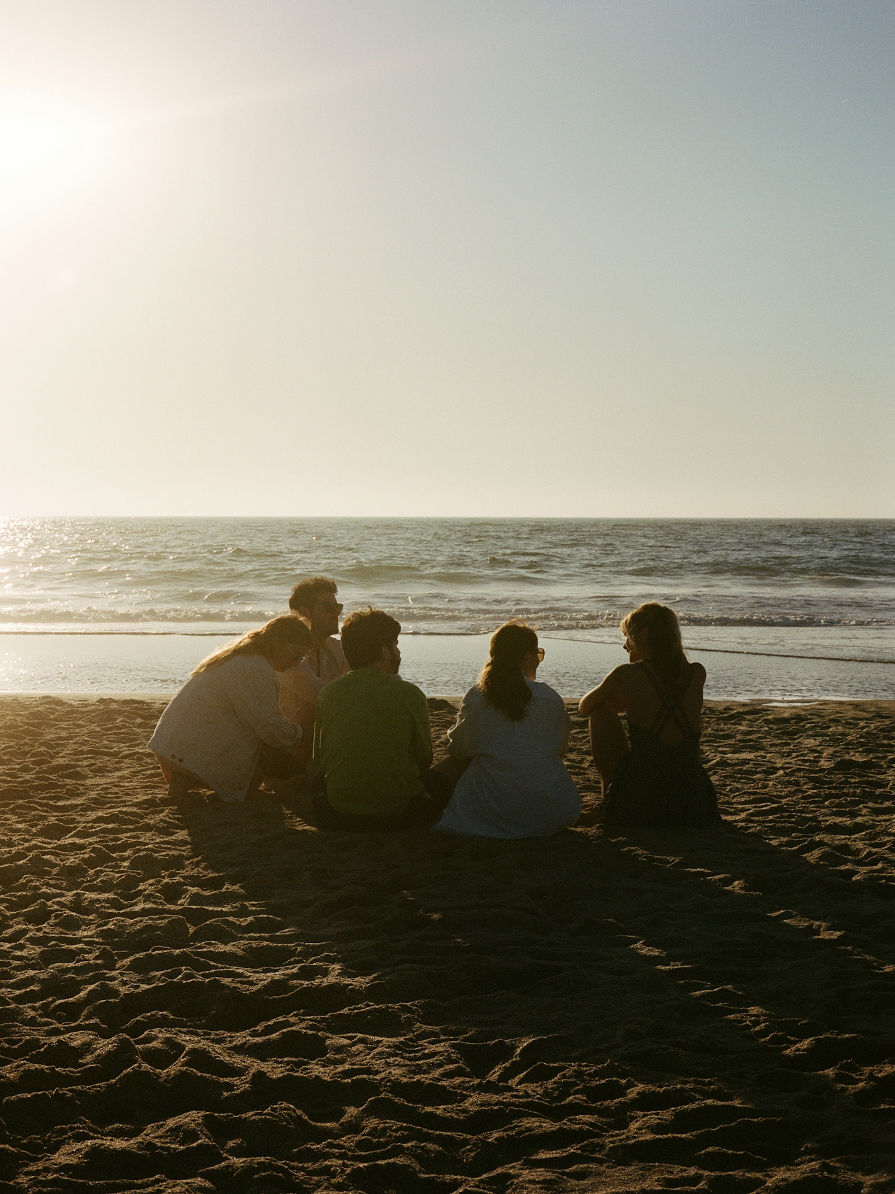 A small group of friends sitting together on a Pacific Northwest beach at golden hour, the ocean glowing behind them.