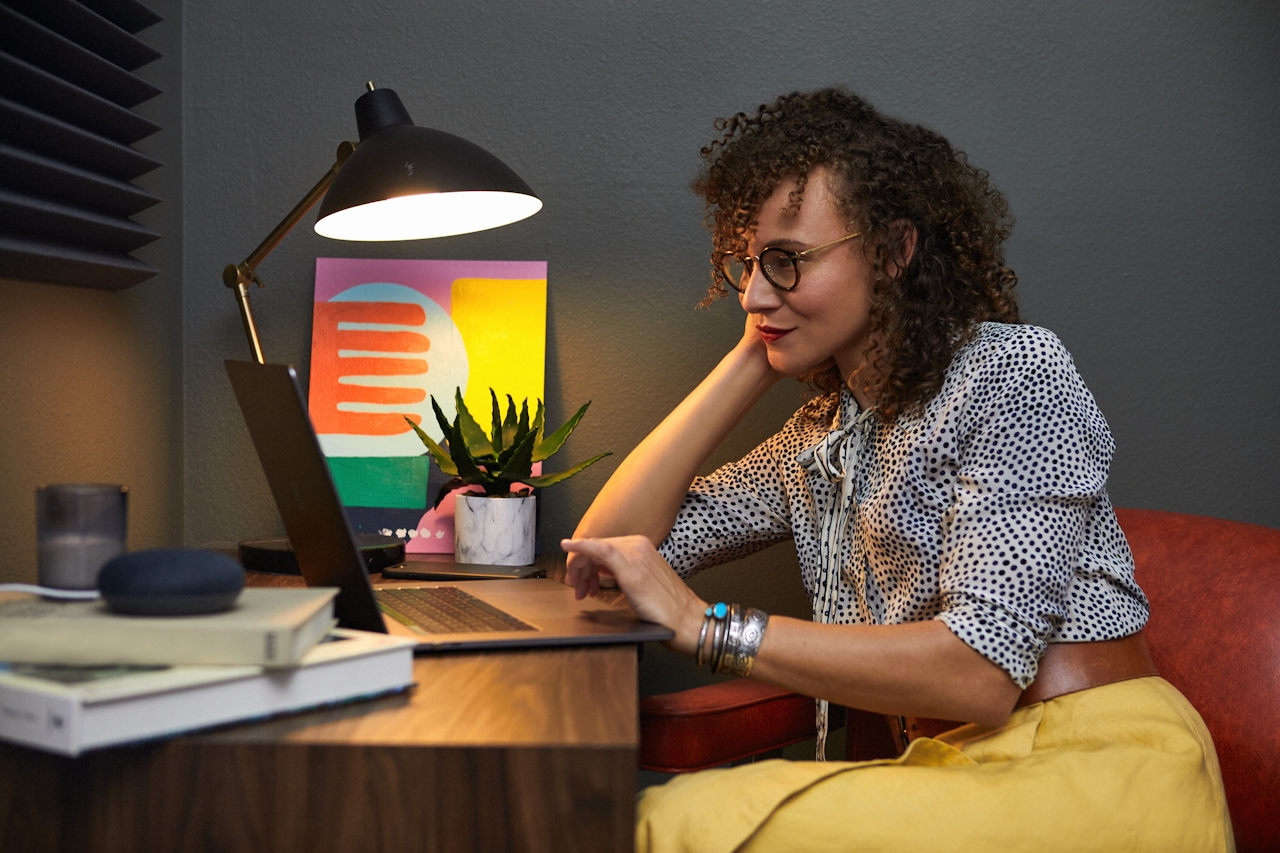 A young woman sitting at a sunlit home desk, working on a laptop with a houseplant and books nearby — a quiet, focused remote-work moment.