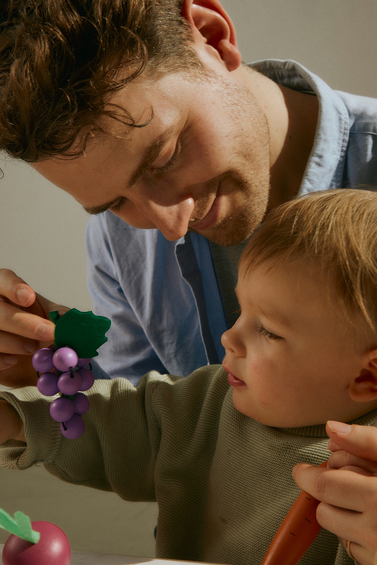A father leaning down to play with his toddler, both focused on a wooden bunch of grapes — a tender, everyday moment of caregiving.