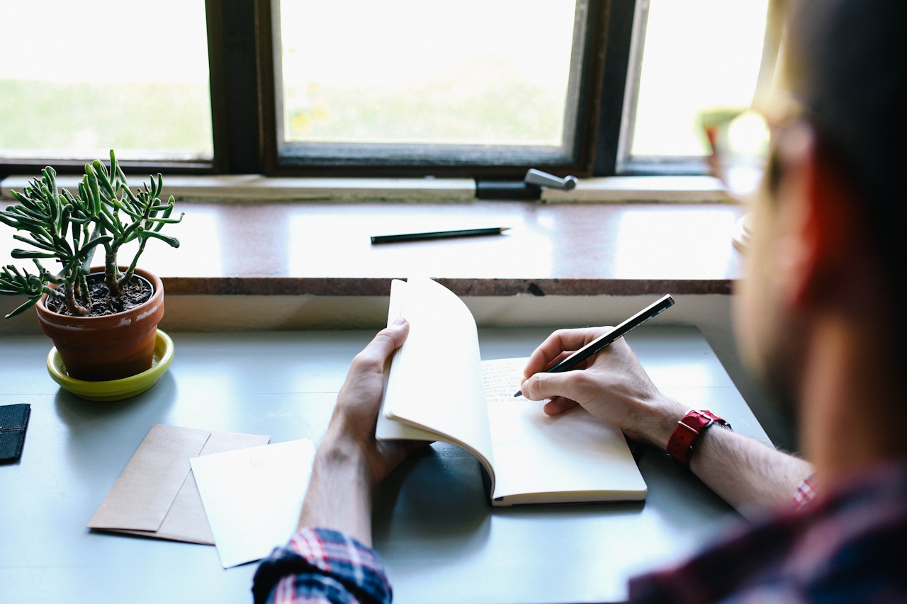A person sitting at a window-side desk, writing in an open notebook with a houseplant nearby — quiet morning ritual.