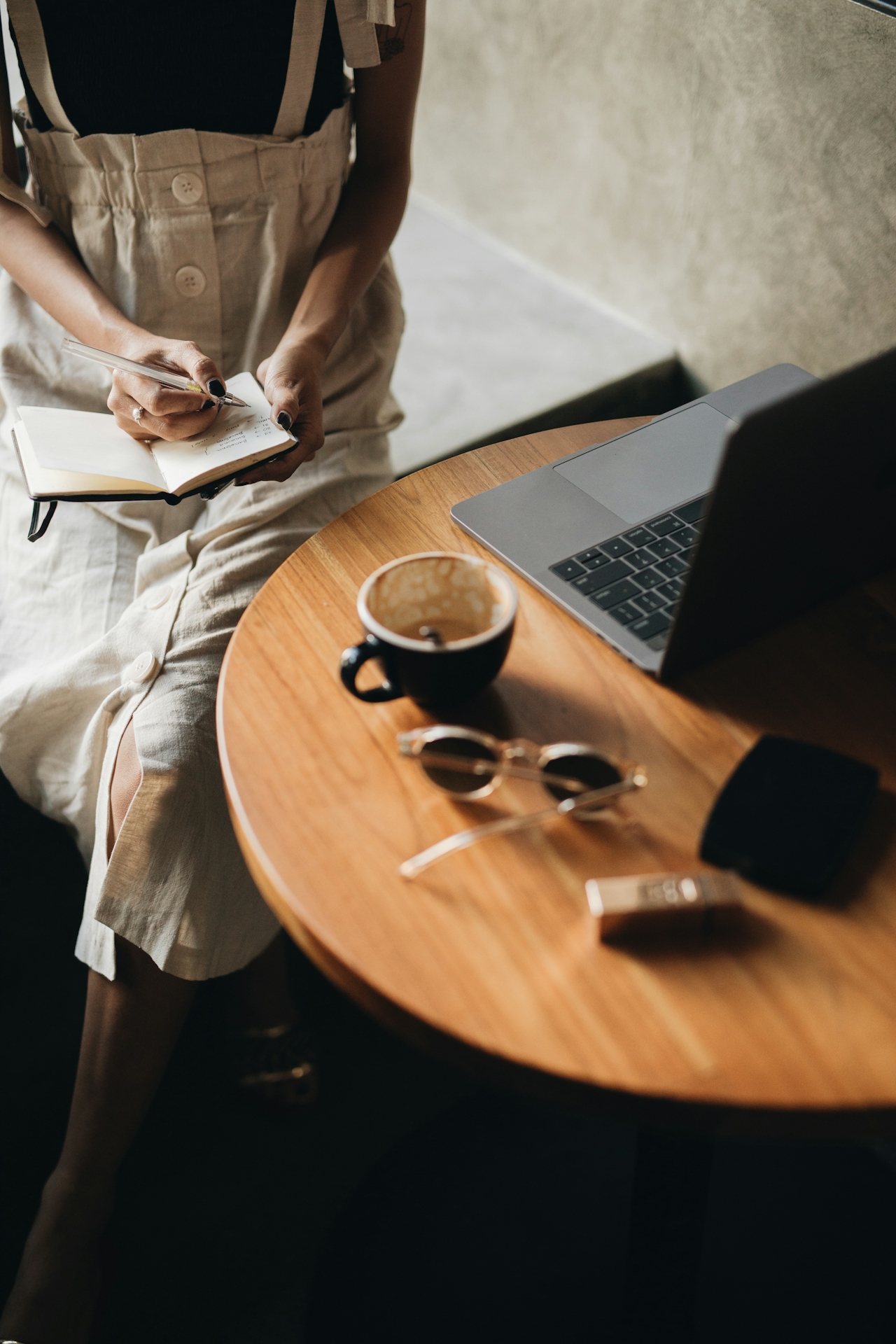 A person sitting with a notebook and pen at a cafe table, laptop nearby, coffee cup and sunglasses on the table — a quiet, unhurried moment of working remotely.