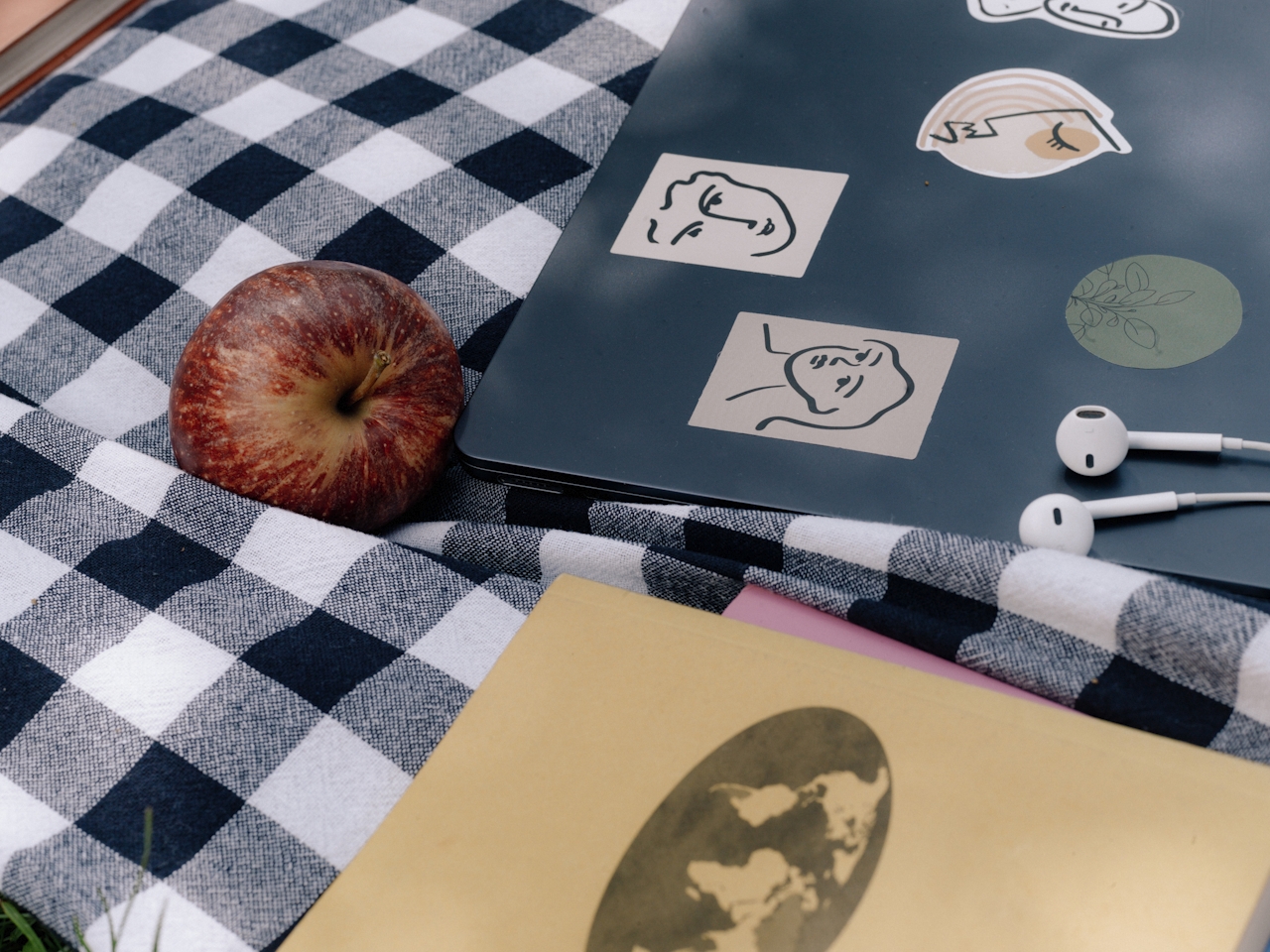 A laptop with hand-drawn stickers, an apple, earbuds, and a book on a gingham picnic blanket — a quiet study setup outdoors.