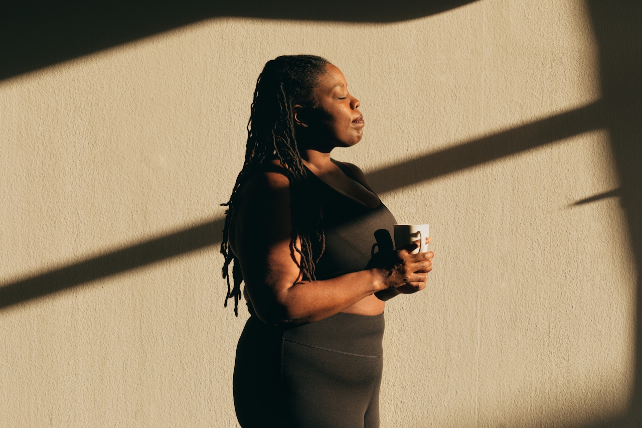 A woman holding a coffee mug, eyes closed in a moment of quiet pause, golden window-light shadows cast across the wall behind her.