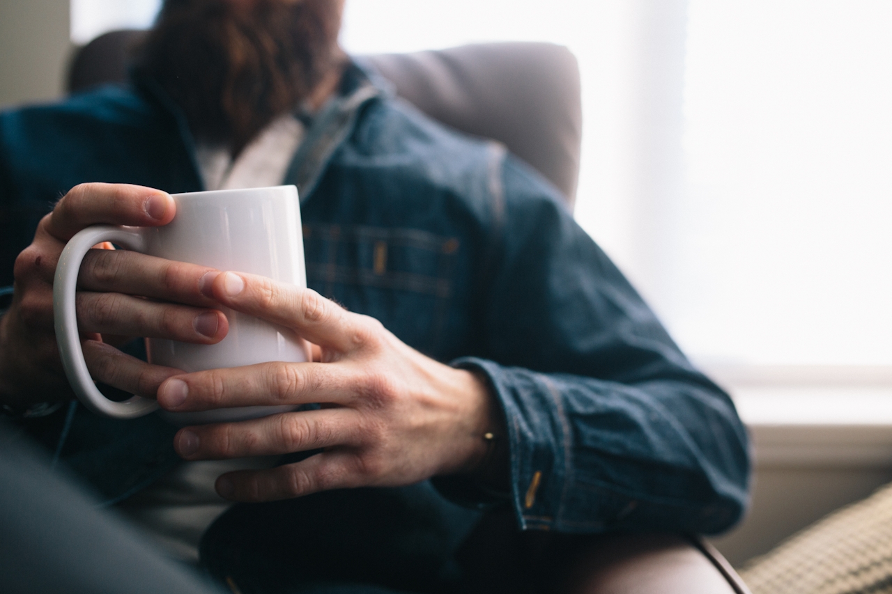 A man relaxing on a couch by a sunlit window, both hands wrapped around a warm mug of coffee — a contemplative pause.