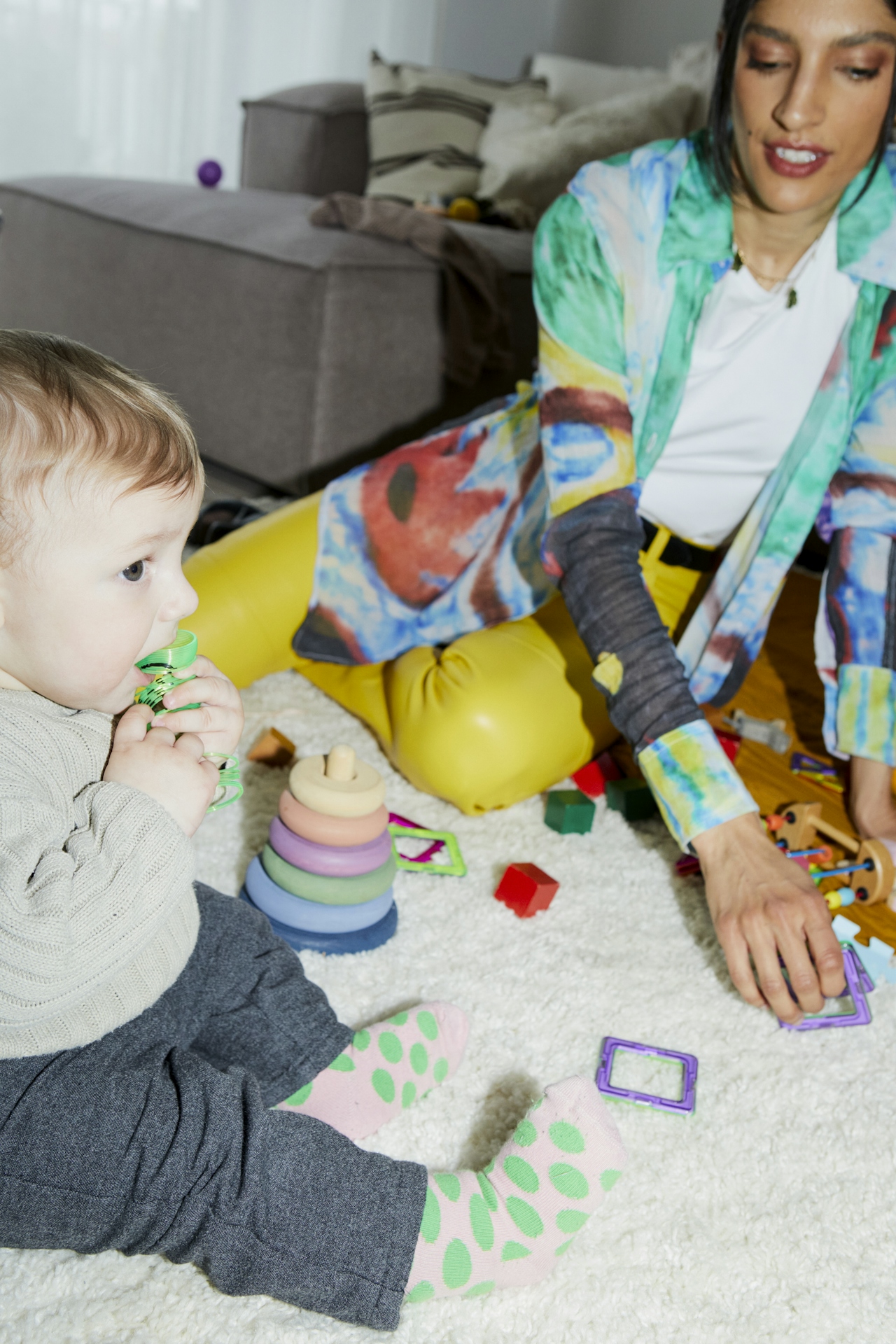 A mother sitting on a living-room rug, playing with her toddler among scattered toys — an unposed moment of everyday parenting.