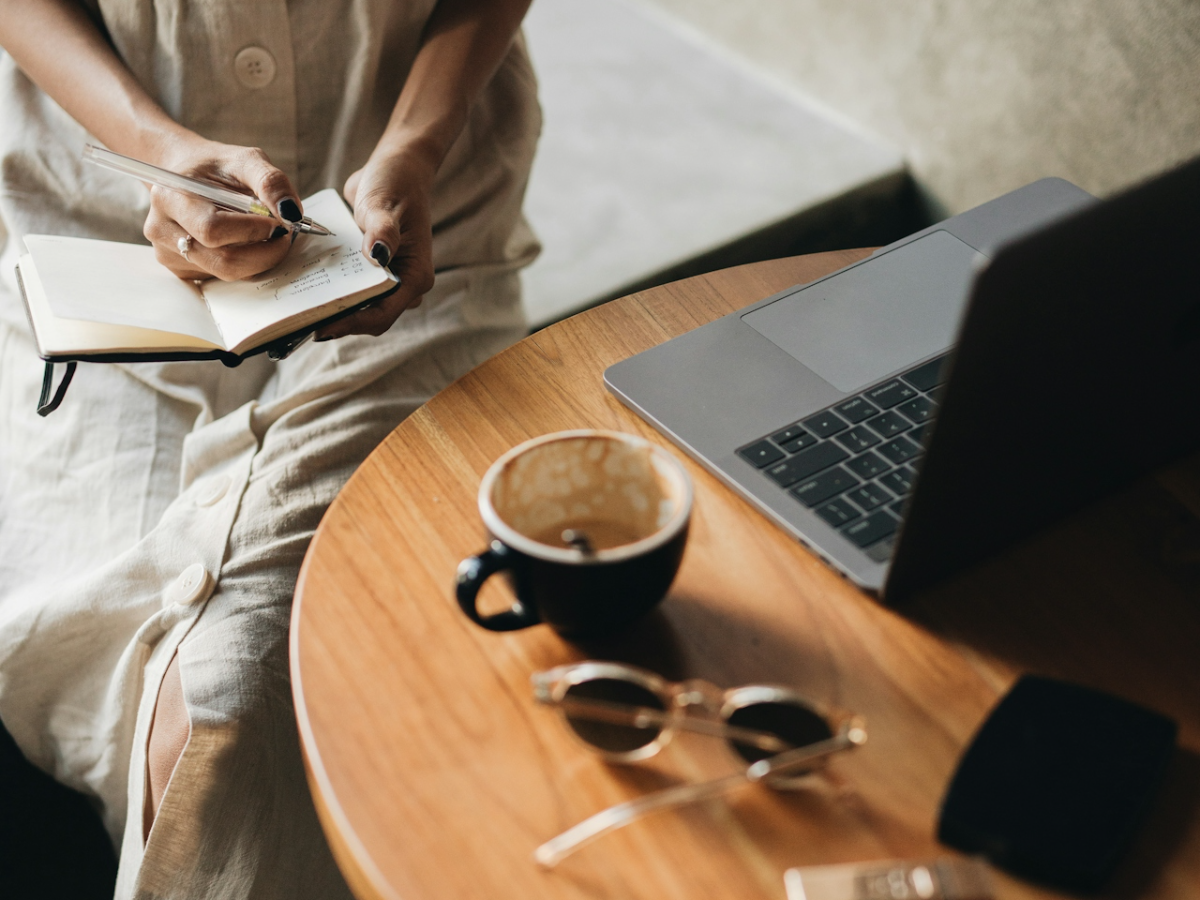A person writing in a notebook at a table beside a laptop and a cup of coffee