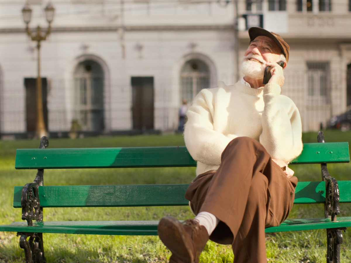 An older adult sitting outdoors on a park bench, talking on a phone in the sun