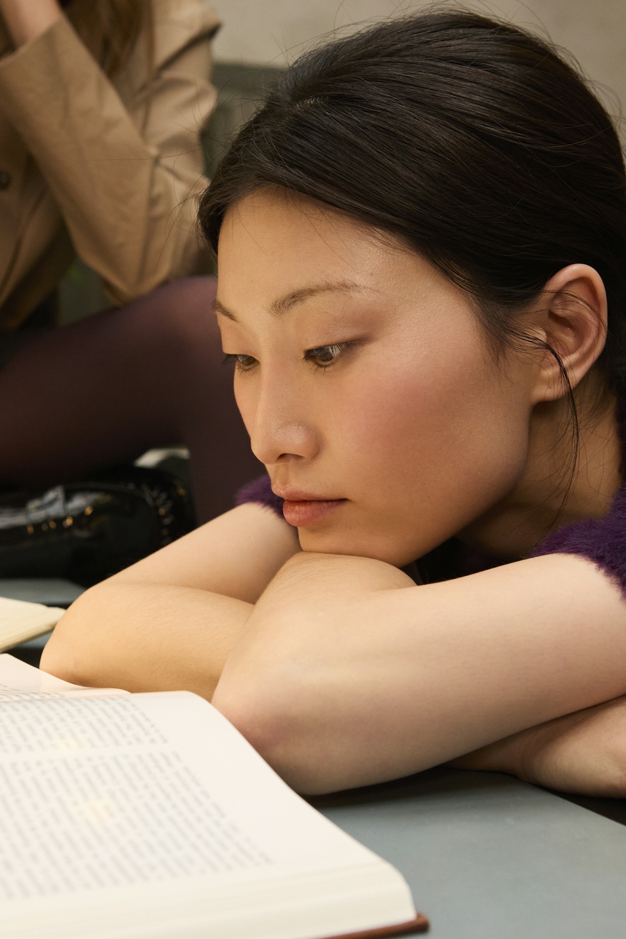A young adult resting her chin on her arms, reading intently from an open book — a quiet moment of focused attention.