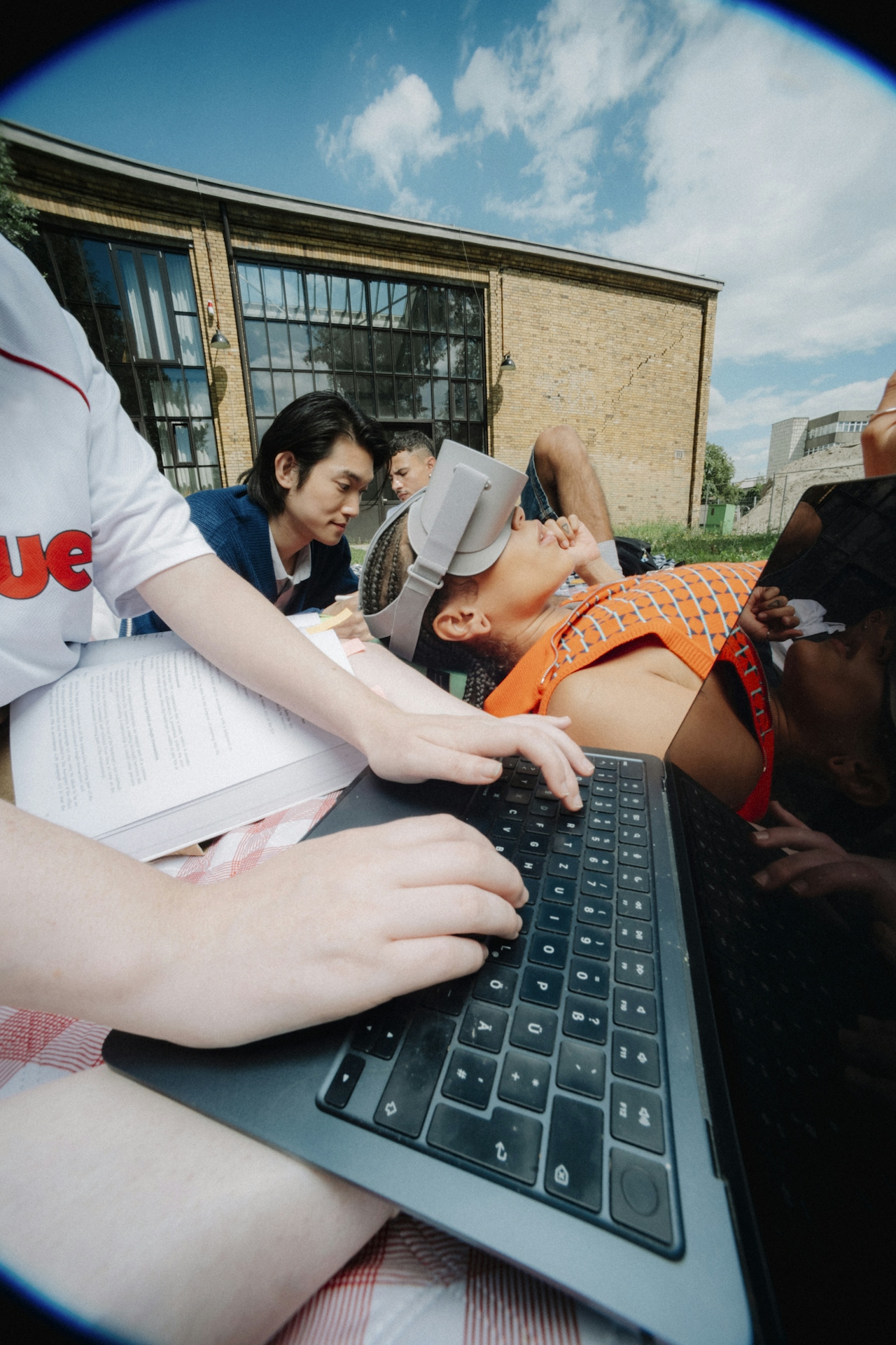 A group of students studying together outdoors on a summer day — laptops, books, and headphones in the late afternoon sun.